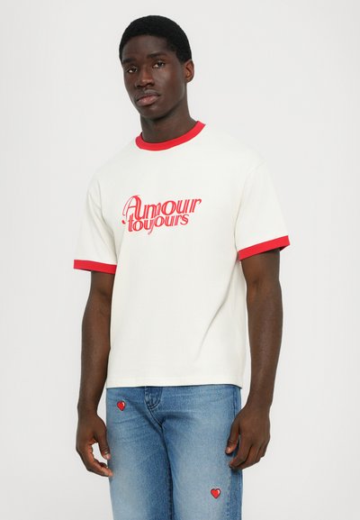 Hvid kortærmet T-shirt med rød krave og manchetter, der har teksten "Amour toujours" i fed rød skrift. Bæres med blå jeans dekoreret med røde hjertepunkter.