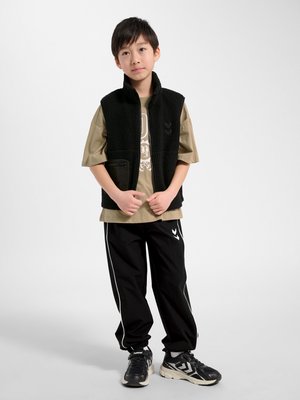 Jongen staat, draagt een zwart fleecevest, een oversized kaki T-shirt, zwarte trainingsbroek met witte strepen en zwarte sneakers.