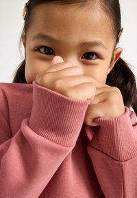 Felpa rosa a maniche lunghe con polsini a coste, che mostra un primo piano di un bambino che copre la bocca con il tessuto, evidenziando la sua morbidezza.