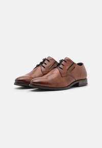 Zapatos de vestir de cuero marrón con un acabado pulido, punta redonda, diseño con cordones y cintas negras contrastantes. Tacón ligeramente elevado.