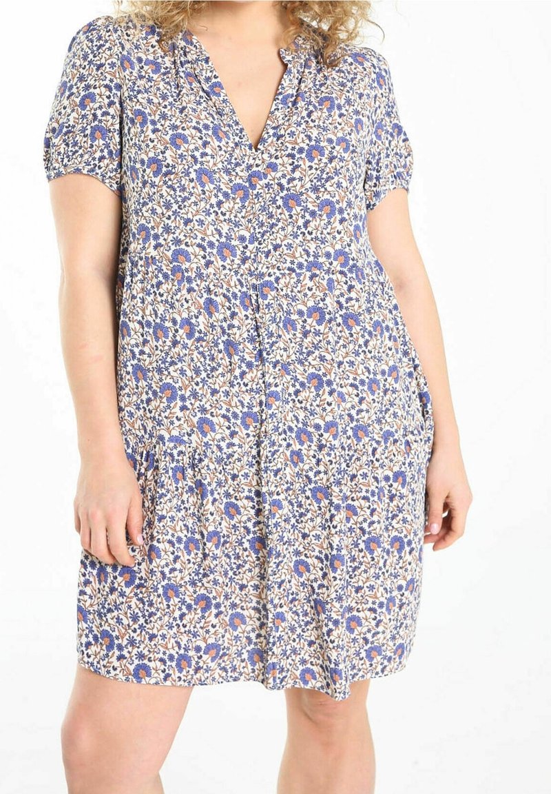 Nainen, jolla on lyhythihainen V-pääntie-mekko, jossa on sinisiä ja oransseja kukkakuoseja valkoisella taustalla.