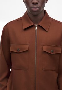 Homme portant une veste marron zippée avec deux poches à rabat à l'avant, fermées par des boutons noirs, et un col pointu sur fond blanc.