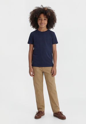 Otrok z kodrastimi lasmi, oblečen v temnomodro majico, bež hlače in rjave čevlje, stoji na enobarvni beli podlagi.