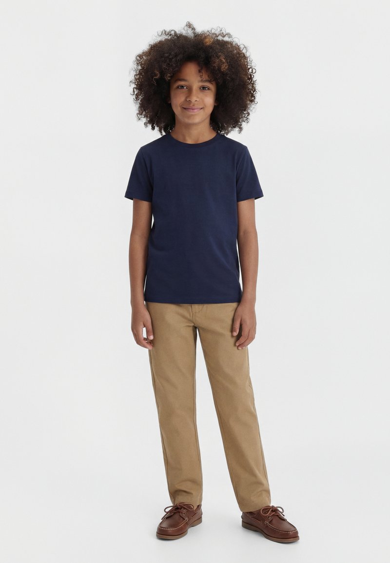 Copil cu păr creț, purtând un tricou albastru închis, pantaloni bej și pantofi maro, stând într-un fundal alb simplu.