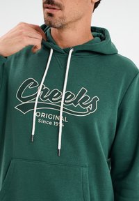 Felpa verde realizzata in morbido tessuto, dotata di una tasca frontale e un cordino bianco. Il design include la scritta "Creeks Original" in bianco.
