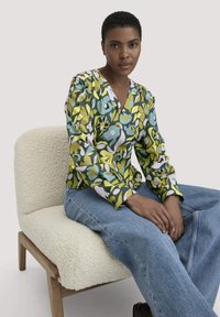 Donna seduta con capelli corti, che indossa una blusa floreale verde e gialla e jeans blu, su una sedia di tessuto crema con texture, davanti a uno sfondo semplice.