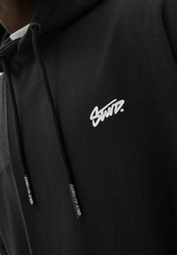 Sweat à capuche noir en tissu doux avec capuche à cordon de serrage. Présente un logo blanc sur la poitrine et des étiquettes de marque sur les cordons.