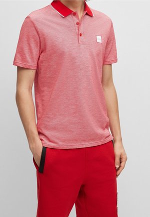 Mann trägt ein kurzärmeliges rotes Poloshirt mit kontrastierendem Kragen und roten Hosen, Hände teilweise in den Taschen, vor einem einfarbigen Hintergrund.