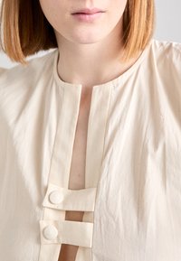 Top de culoare crem, cu guler înalt, fante în față și două închideri cu butoane rotunde. Materialul are o textură netedă, ușor șifonată.