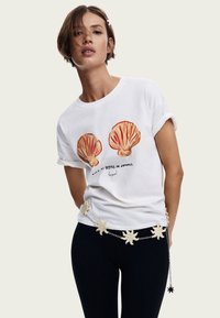 Hvid bomulds T-shirt med to orange muslingeskaller grafik, med teksten "Livet er bedre om sommeren." Parret med sorte bukser og dekorativ kædebælte.