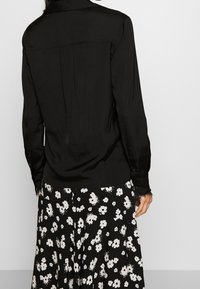 Persona con una blusa negra de manga larga y puños abotonados, y una falda negra con patrones florales blancos, vista desde atrás.