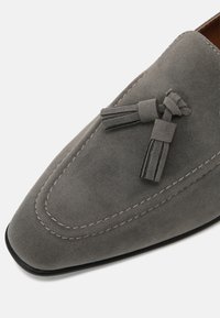 Mocassin gris en daim avec des détails cousus et deux pampilles en daim sur le dessus, présenté en vue rapprochée sous un angle.
