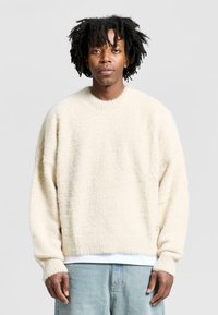 Cream-colored fuzzy sweater with a round neck, dropped shoulders, and ribbed cuffs, layered over a white t-shirt, featuring a relaxed fit.