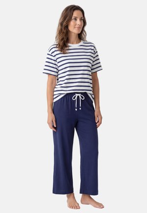 Vrouw staat blootsvoets, gekleed in een wit shirt met navyblauwe strepen en korte mouwen en een navyblauwe broek met trekkoord, tegen een effen achtergrond.