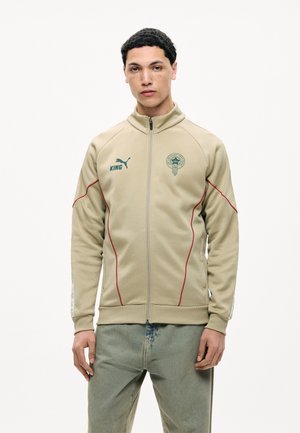 Jeune homme portant une veste beige zippée avec un liseré rouge, le logo Puma et le texte "KING", associé à un jean bleu délavé, debout devant un fond uni.