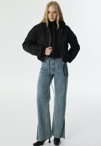 Zwarte cropped bomberjack met zilveren rits, gecombineerd met lichtblauwe wijde jeans met zijsplitten en een donkere top eronder.