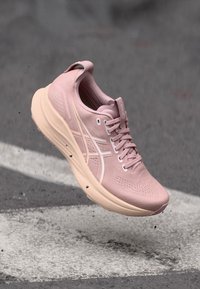 Pink athletic shoe with a textured upper, rounded toe, cushioned sole, and white accents. Features a lace-up design and logo detailing.