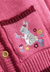 Gestrickte rosa Strickjacke aus strukturiertem Stoff, mit einer aufgestickten grauen Häschen und bunten Blumenmustern auf einer vorderen Tasche. Braunes Knopfdetail.