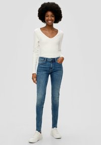 Uske skinny traperice u srednje plavom denimu, s visokim strukom, pet džepova i suptilnim izbljeđivanjem. Upareno s bijelom majicom dugih rukava i bijelim tenisicama.