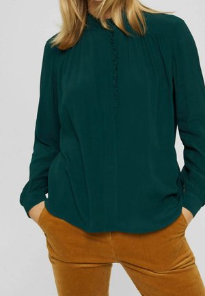 Kobieta ubrana w ciemnozieloną bluzkę z długim rękawem ozdobioną falbankami oraz musztardowe sztruksowe spodnie, z rękami w kieszeniach.