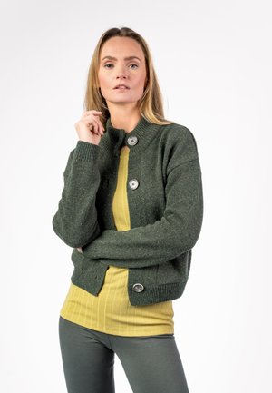 Frau mit langen blonden Haaren, die eine grüne, geknöpfte Strickjacke über einem gelben, gerippten Oberteil und grauen Hosen trägt, steht mit einer Hand in der Nähe ihres Gesichts.