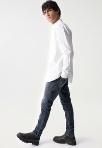 Camicia bianca a maniche lunghe con colletto abbottonato, abbinata a jeans slim-fit blu scuro e stivali neri con suola in gomma. Sfondo neutro.