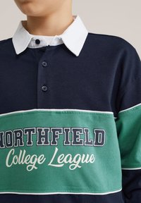 Laivastonsininen ja vihreä pitkähihaisen polopaidan jossa valkoinen kaulus ja napit, ja rinnassa teksti "NORTHFIELD College League".