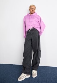 Person with short blonde hair wearing a purple hoodie, loose black cargo pants, and leopard print sneakers standing on blue carpet.