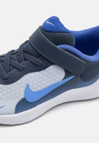 Chaussure de sport bleue et grise avec tige en mesh, languette rembourrée et sangle Velcro. Dispose d'une semelle blanche et du logo Nike en bleu.