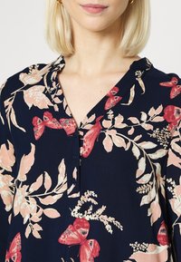 Blusa floreale navy con motivi di fiori rossi e rosa e farfalle, con pattina con bottoni e una texture di tessuto morbido.