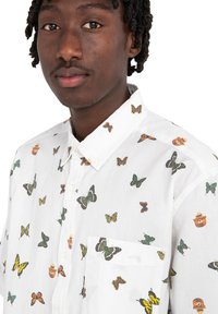 Camisa blanca de botones con estampados variados de mariposas y animales. Presenta un bolsillo y una textura suave y ligera.