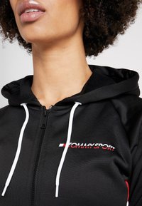 Primo piano di una persona che indossa una felpa nera con zip, con lacci bianchi e il logo rosso e bianco di Tommy Sport sul petto.