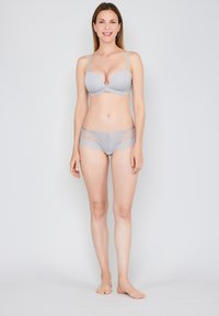 Femme souriante debout pieds nus, portant un soutien-gorge en dentelle gris clair et un slip hipster assorti, devant un fond blanc uni.