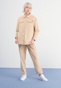 Beige kordfløyelsjakke med to brystlommer, sammen med matchende bukser. Hvite sneakers fullfører antrekket mot en ensfarget bakgrunn.