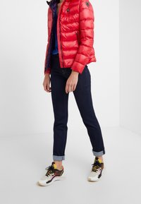 Rode gewatteerde jas met ritsen, hoge kraag en logo-patch; blauw overhemd eronder, donkere spijkerbroek met opgerolde pijpen en meerkleurige sneakers.