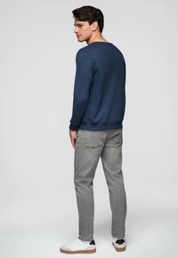 Mand set fra siden iført navyblå sweatshirt, grå jeans og hvide sneakers på en ensfarvet lysegrå baggrund.