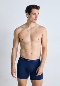 Boxer-briefs bleus en tissu extensible, avec une ceinture de marque portant l'inscription "TOMMY HILFIGER" en lettres blanches. Le modèle se tient devant un fond clair.