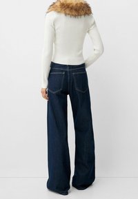 Vit ribbade långärmad topp med en fuskpälskrage, kombinerad med mörkblå vida denimjeans som har bakfickor och kontrasterande sömmar.