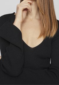 Czarny sweter z prążkowanej dzianiny z głębokim dekoltem w kształcie V i rozszerzanymi rękawami. Teksturowany materiał o gładkim wykończeniu. Zbliżenie na dekolt i rękawy.