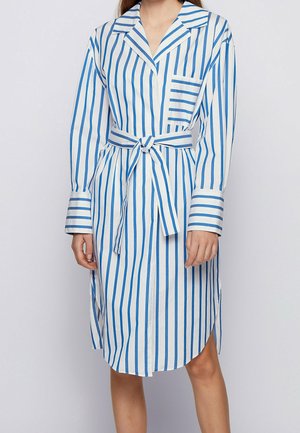 Femme portant une robe blanche mi-longue à rayures verticales bleues, avec une poche poitrine, des manches longues et une ceinture nouée à la taille.