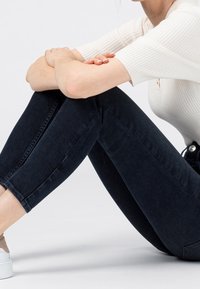 Jeans ajustados de color azul oscuro con detalles descoloridos, combinados con una camiseta ligera de canalé. La persona está sentada con los brazos apoyados en las rodillas, usando zapatillas ligeras.