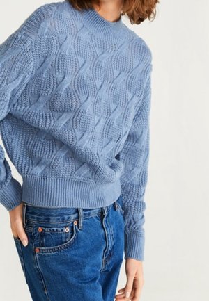 Kobieta w jasnoniebieskim swetrze w warkocz, wsadzonym w wysoki stan niebieskich dżinsów, stojąca na tle jednolitego białego tła.