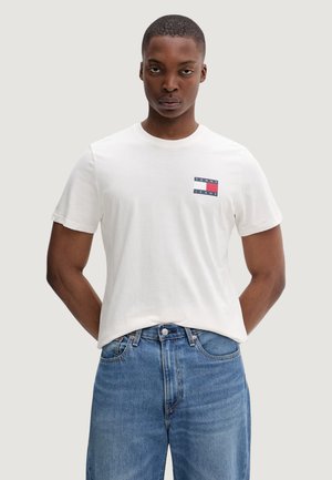 Hombre de pie con las manos detrás de la espalda, vistiendo una camiseta blanca de Tommy Jeans y jeans azules sobre un fondo claro y liso.