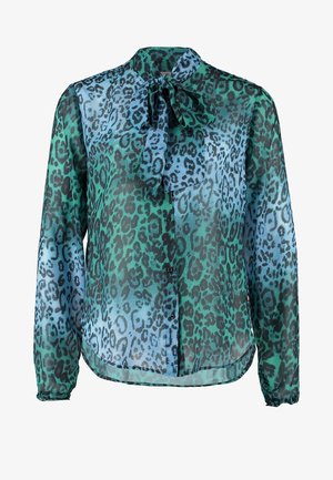 Blouse transparente à imprimé léopard en vert et bleu, avec un col noué. Associée à un pantalon blanc ajusté et des chaussures claires. Tenant une pochette verte.