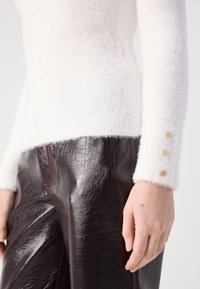 Weißer, flauschiger Strickpullover mit drei Holzknöpfen an den Ärmeln, kombiniert mit glänzenden, dunkelbraunen Lederhosen, die rote Nähte aufweisen.