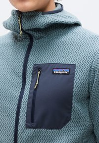 Άτομο που φοράει μια μπλε-πράσινη υφασμάτινη φλις ζακέτα με μια σκούρα μπλε τσέπη στο στήθος με φερμουάρ, η οποία διαθέτει ένα λογότυπο της Patagonia.