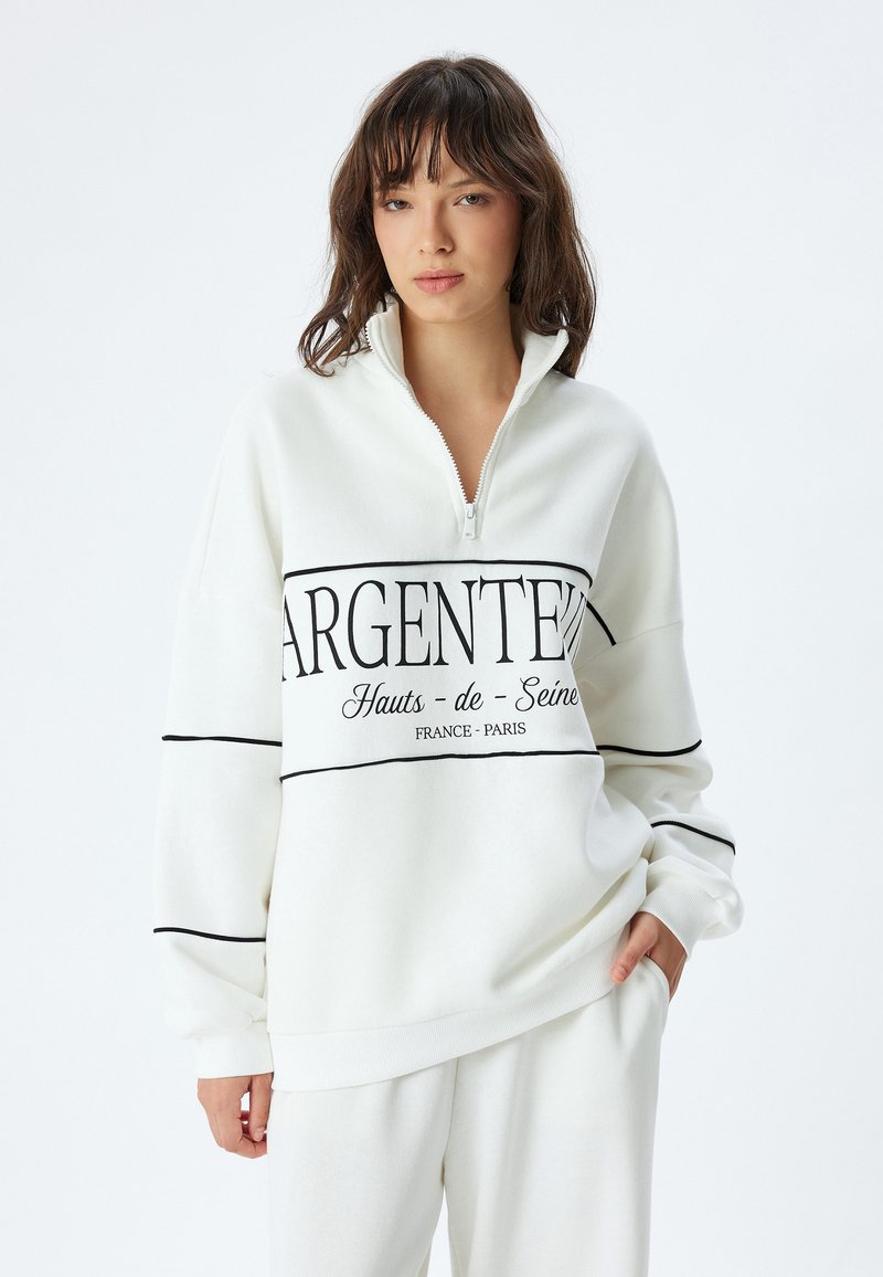 Sudadera de forro polar blanca con media cremallera, que presenta texto y líneas en negro. Corte relajado con puños y dobladillo acanalados para mayor textura.