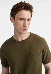 Oliwkowy, krótki rękaw T-shirt wykonany z teksturowanej tkaniny. Posiada okrągły dekolt oraz kieszeń na lewej piersi z detalem metki.