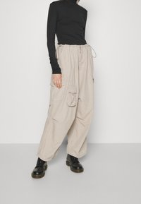 Beige Cargo-Hosen mit großen Seitentaschen, Kordelzugbund und einer lässigen Passform, kombiniert mit einem eng anliegenden schwarzen Langarmoberteil und schwarzen Stiefeln.