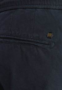 Donkerblauwe stoffen broek met een gestructureerde afwerking, voorzien van een achterzak met een hexagonaal zwart metalen accent en subtiele stikdetails.